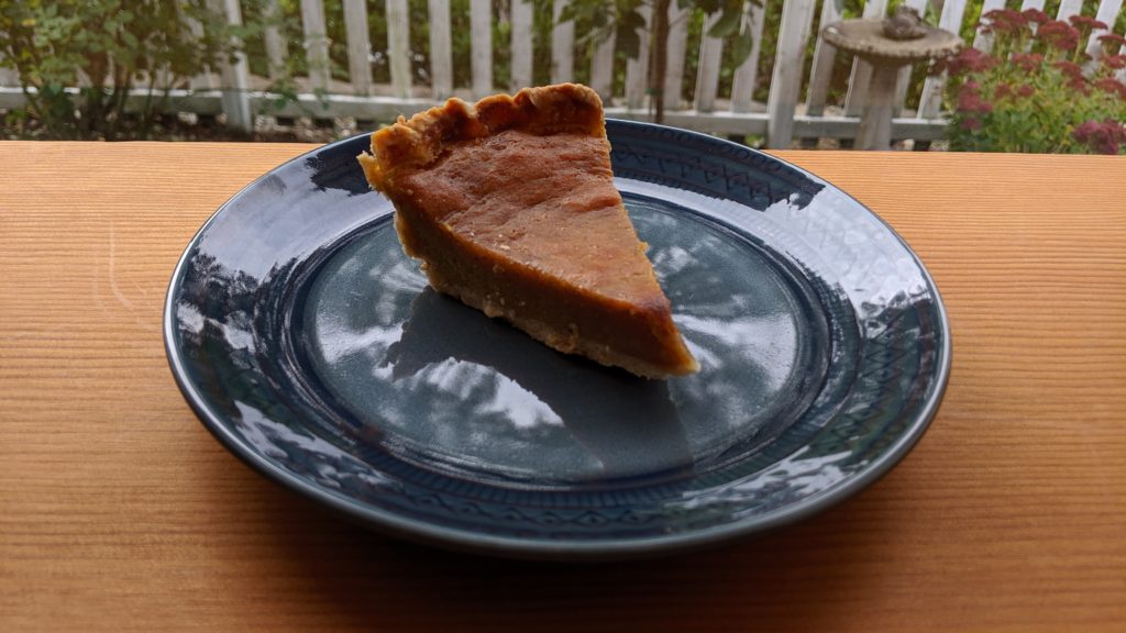 A slice of fresh pumpkin pie on a navy blue plate on a vertical-grain Douglas Fir table with a view of a white picket fence out the window.